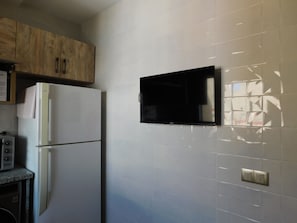 Une cuisine avec un réfrigérateur blanc, des armoires en bois, un téléviseur noir fixé au mur et un four à micro-ondes posé sur le plan de travail.