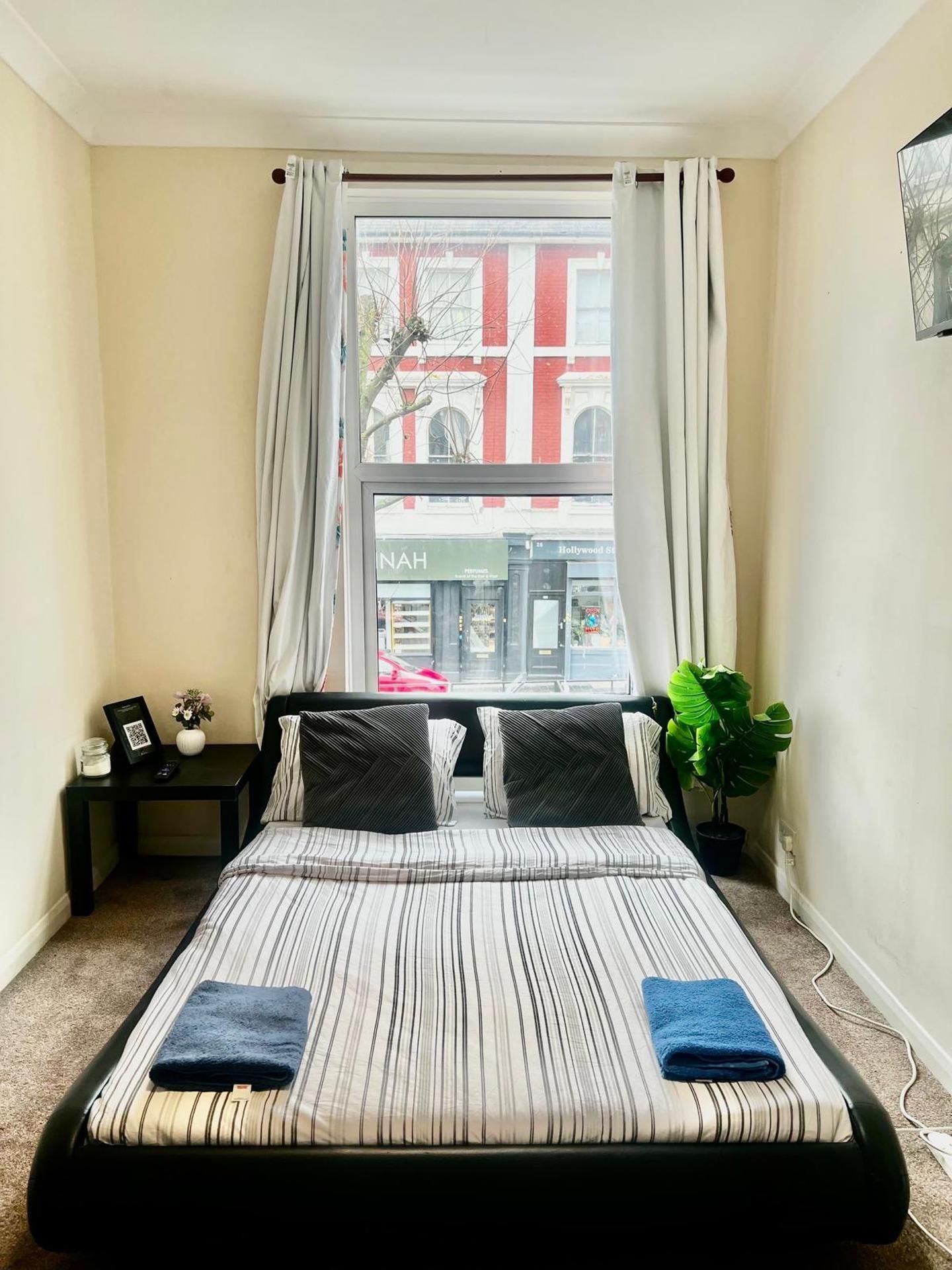A bedroom with a bed, a window with curtains, a small table with a vase and a picture frame, and a potted plant.