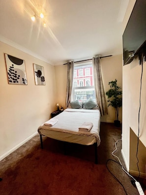 A hotel room with a bed, a window with curtains, a plant, and artwork on the wall. - Apartment Sleeps 4 Near Station (London)