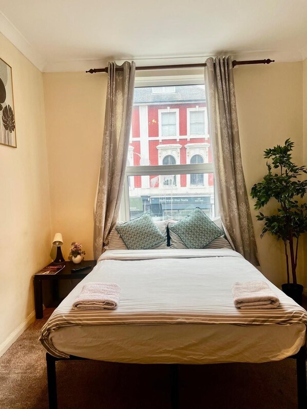 A hotel room with a bed, a nightstand, a lamp, a potted plant, and a window with curtains. - Apartment Sleeps 4 Near Station (London)