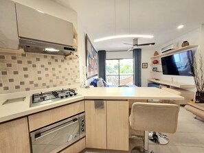 Cozinha moderna com fogão a gás, forno e micro-ondas embutidos. Há uma ilha grande com uma cadeira e uma TV de tela plana fixada na parede.