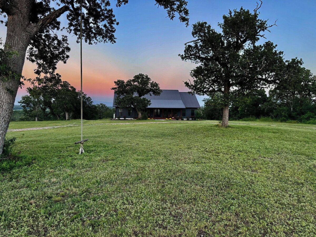 Um campo gramado com uma casa e árvores.