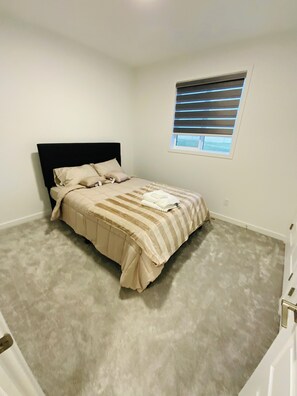 A bedroom with a bed, a window with blinds, and a door. - Bright, Spacious & Peaceful | Modern 3BR West Lethbridge Retreat (Lethbridge)