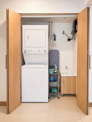 A compact laundry room with a washing machine, ironing board, and a sink. - Stay Longer in Jacó - Beach 100m - Pool - Gym (Jacó)