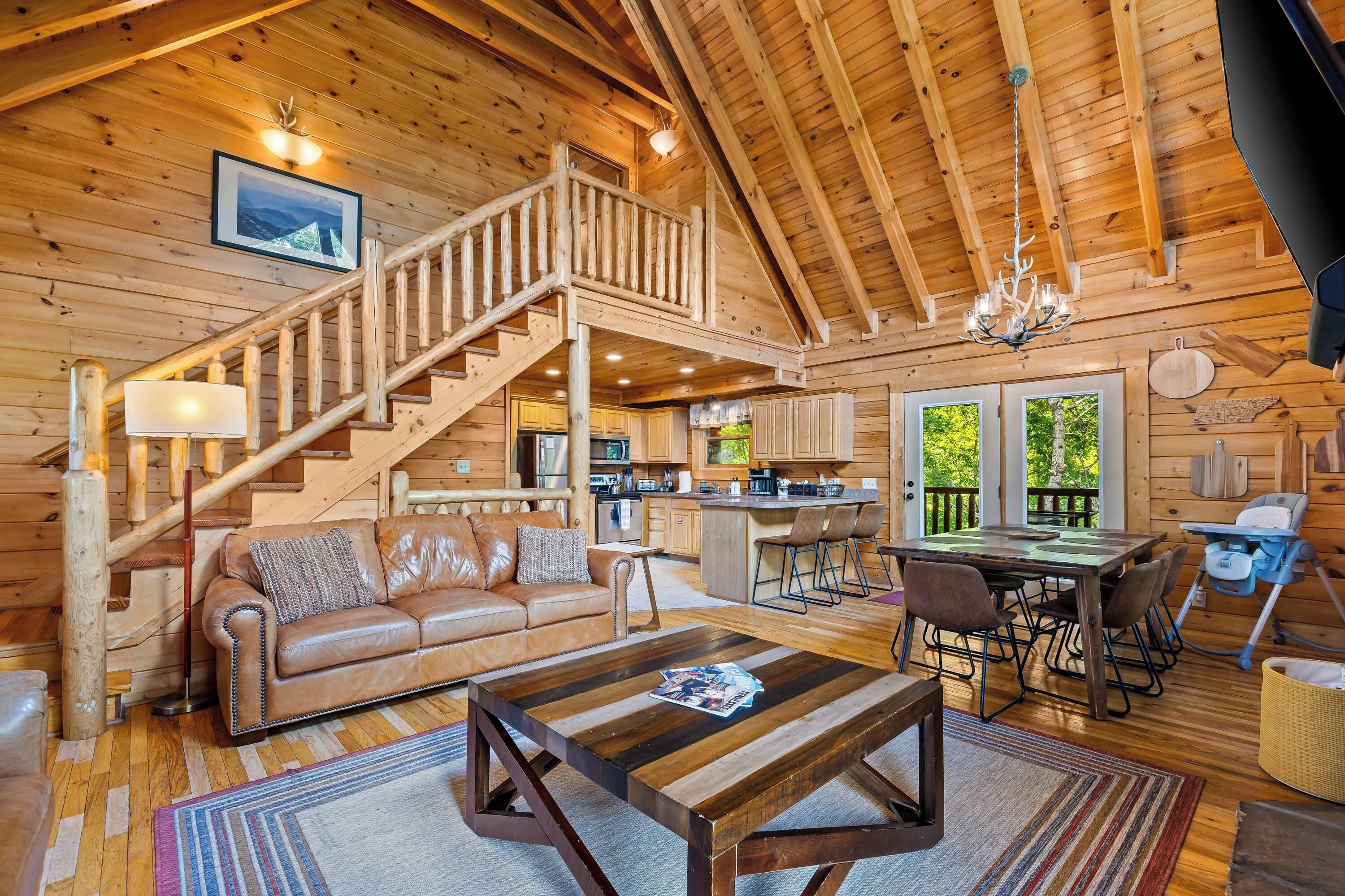 A log cabin interior with a wooden staircase, a leather sofa, a dining table with chairs, and a kitchen area.
