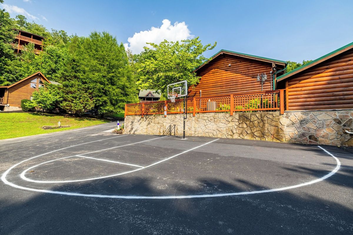 Un terrain de basket-ball entouré d’une clôture en bois et d’un mur de pierre, bordé d’arbres et adjacent à un bâtiment doté d’un toit végétalisé.