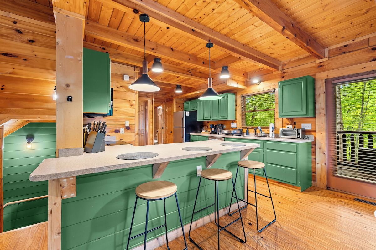 A wooden cabin interior with a kitchen island, bar stools, and a large window with a view of greenery.