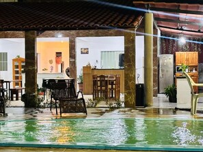 A covered outdoor area with a pool, seating, and a kitchen area visible in the background. - Pousada Costa do Sol  (Porto Seguro)