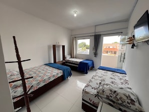 A dormitory room with three beds, a wardrobe, a window with curtains, and a television on the wall.