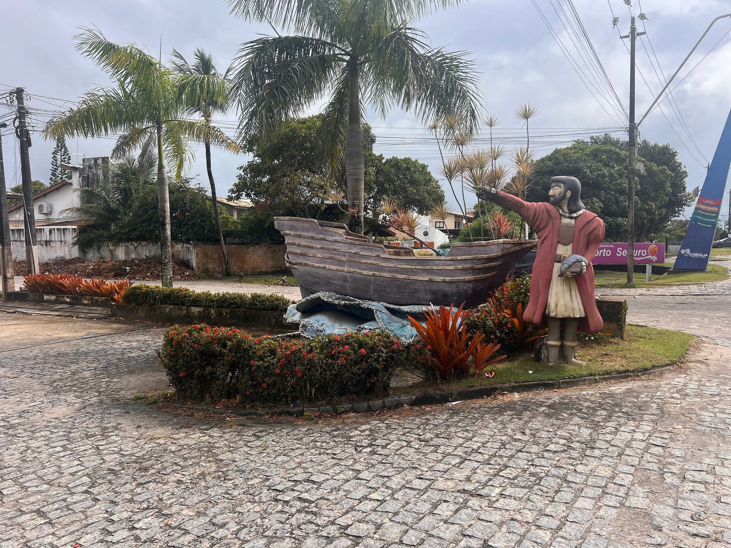 Estátua de um homem segurando um barco, cercada por plantas tropicais e um caminho de pedras.