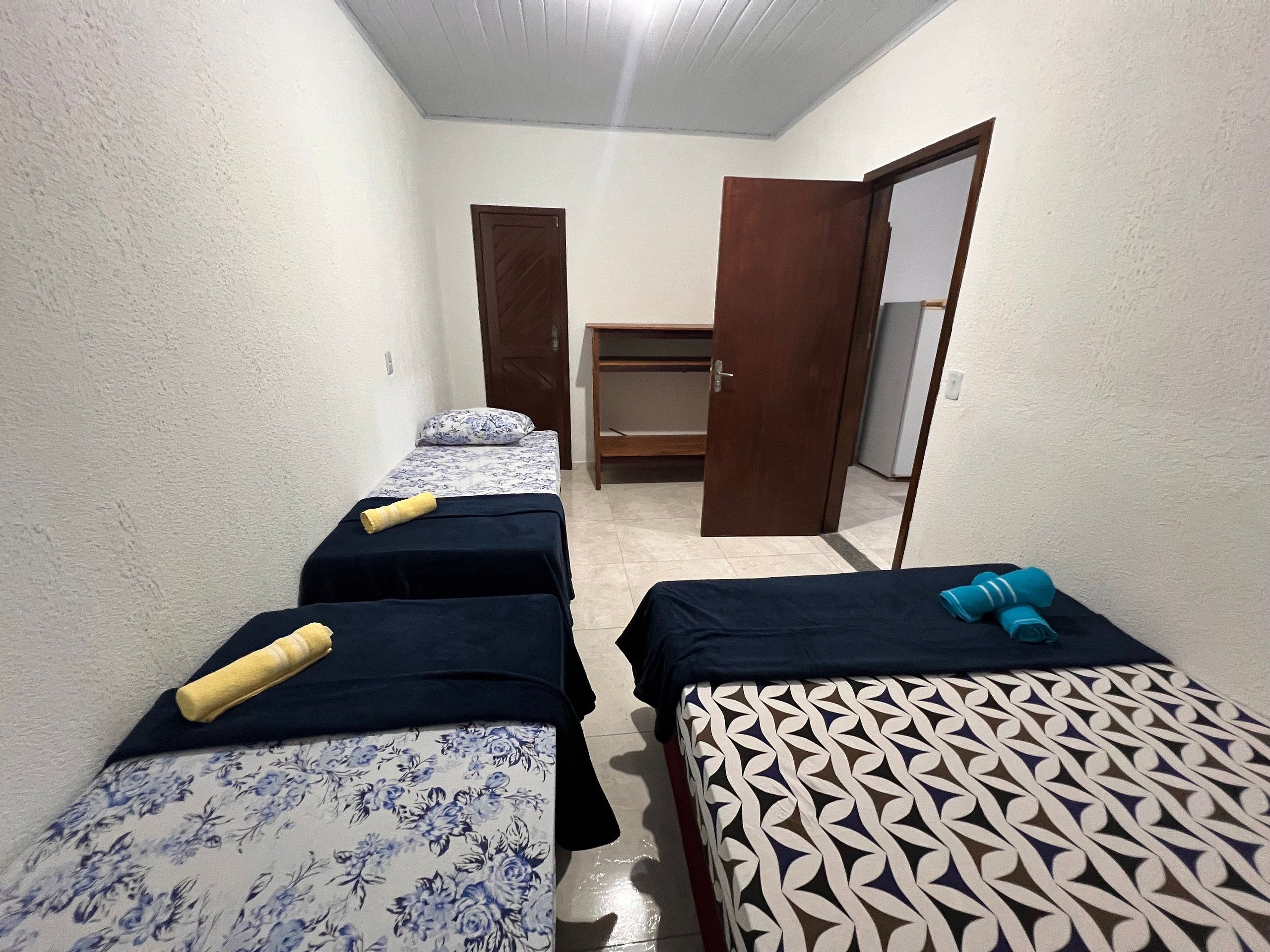 A narrow room with two beds, one with a floral bedspread and the other with a geometric pattern. There is a wooden bench against the wall and a door leading to another room.