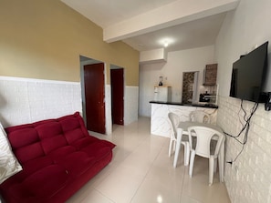 A living area with a red sofa, a dining table with chairs, a kitchen area with a refrigerator, and a TV mounted on the wall.