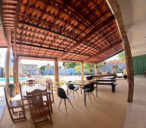 Un patio techado al aire libre con mesas y sillas de madera, una piscina al fondo y una pared azul.