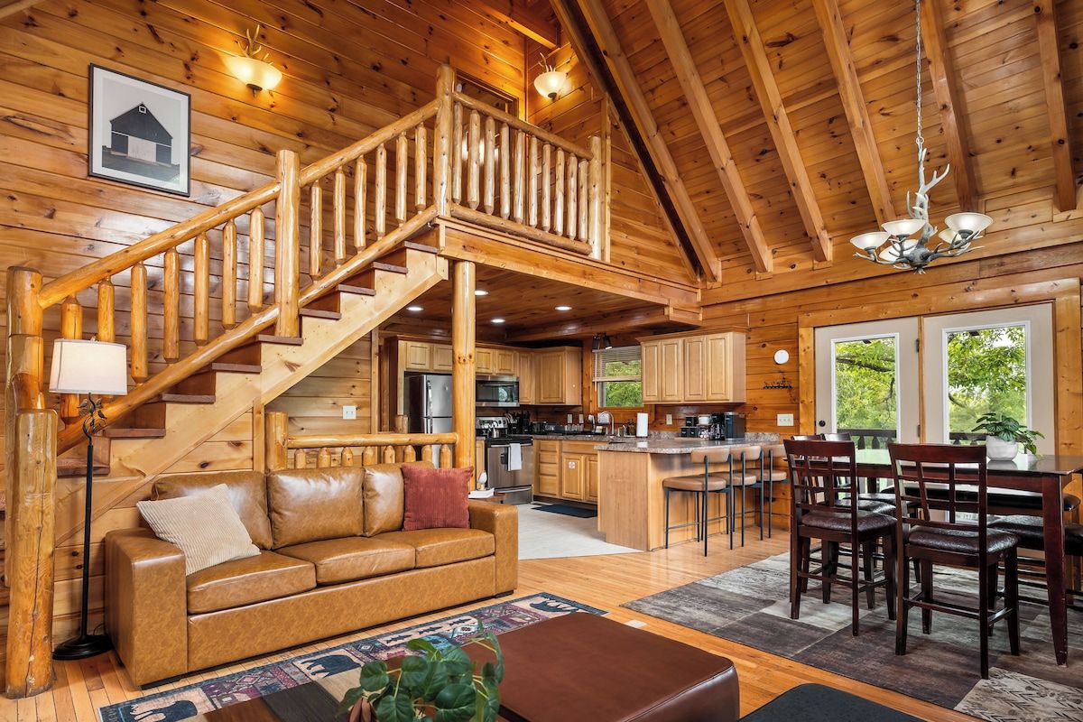 Interior de una cabaña de madera con escalera, cocina, comedor y sala de estar con sofá y mesa de centro.