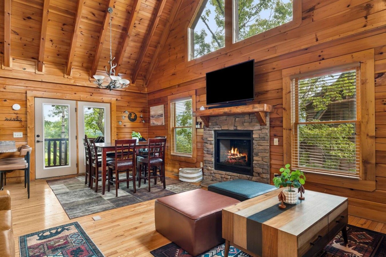 A log cabin interior with a fireplace, dining area, and large windows.