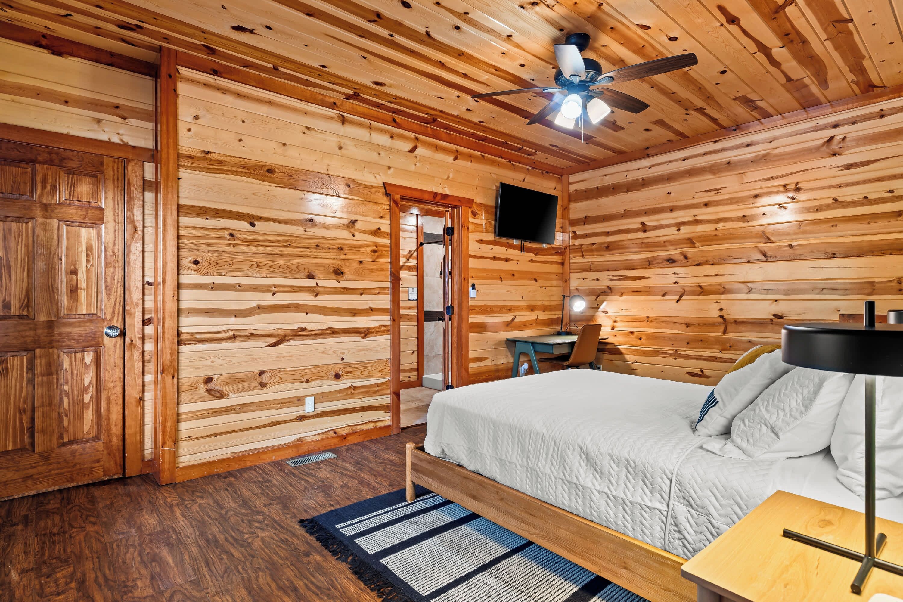 A wooden-paneled room with a bed, a desk, a chair, a television, and a ceiling fan.