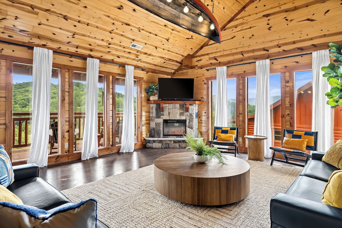 A spacious living room with a fireplace, wooden ceiling, and large windows.