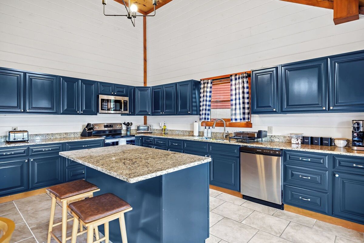 A kitchen with dark blue cabinets, a granite countertop, a stainless steel dishwasher, and a checkered window valance.