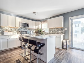A modern kitchen with white cabinets, a central island, black bar stools, a microwave, and a tiled backsplash.