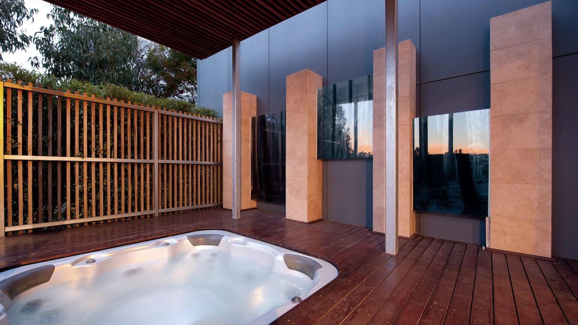 Un área moderna de hidromasaje al aire libre con una terraza de madera, una valla metálica y un edificio con grandes ventanales.