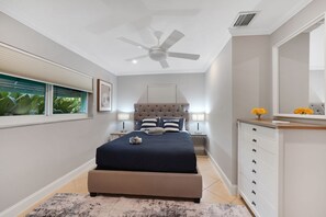 Ein Schlafzimmer mit Bett, Nachttischen, Deckenventilator, Fenster mit Jalousien und einer Kommode mit Schubladen.
