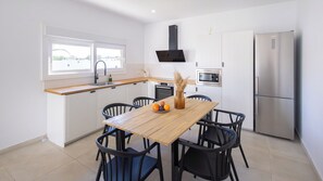 A modern kitchen with white cabinetry, a wooden dining table, black chairs, a built-in oven, and a stainless steel refrigerator. - Casa Maricarmen Conil de la Frontera by Ruralidays (Cádiz)