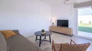 Ein modernes Wohnzimmer mit einer Couch, einem kleinen runden Tisch mit einer Pflanze, einem Fernseher auf einer Holzkommode und Blick auf eine grüne Außenanlage durch eine Glastür.