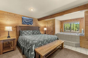 A wooden-paneled bedroom with a bed, two bedside lamps, a bench, a large window, and a bathtub. - Alpine Pension ~ Riverfront, Hot tub, Giant Game Room ~ 1 mile from town! (Leavenworth)