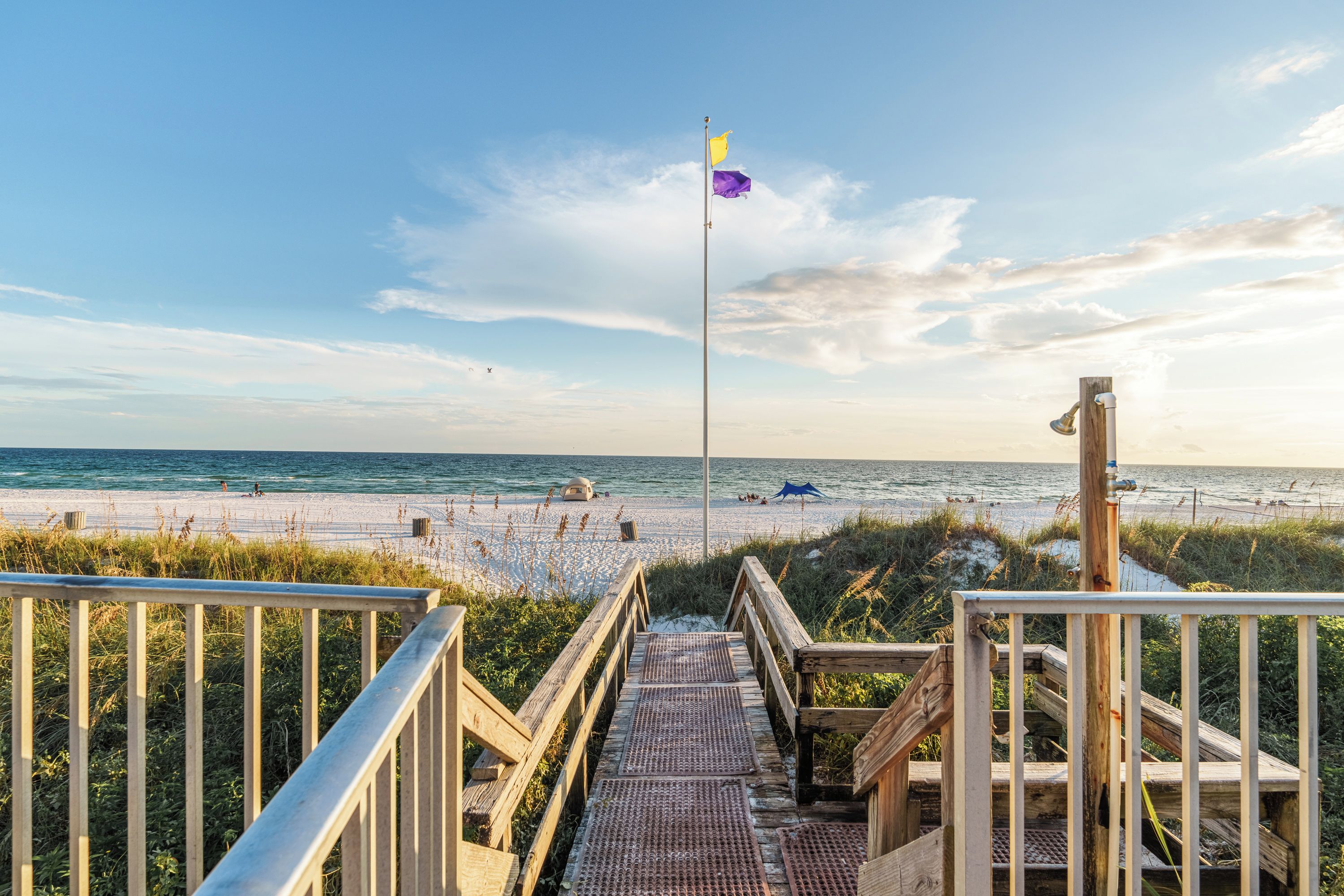 Ein Holzsteg führt zu einem Strand mit einem Fahnenmast und einer Flagge.
