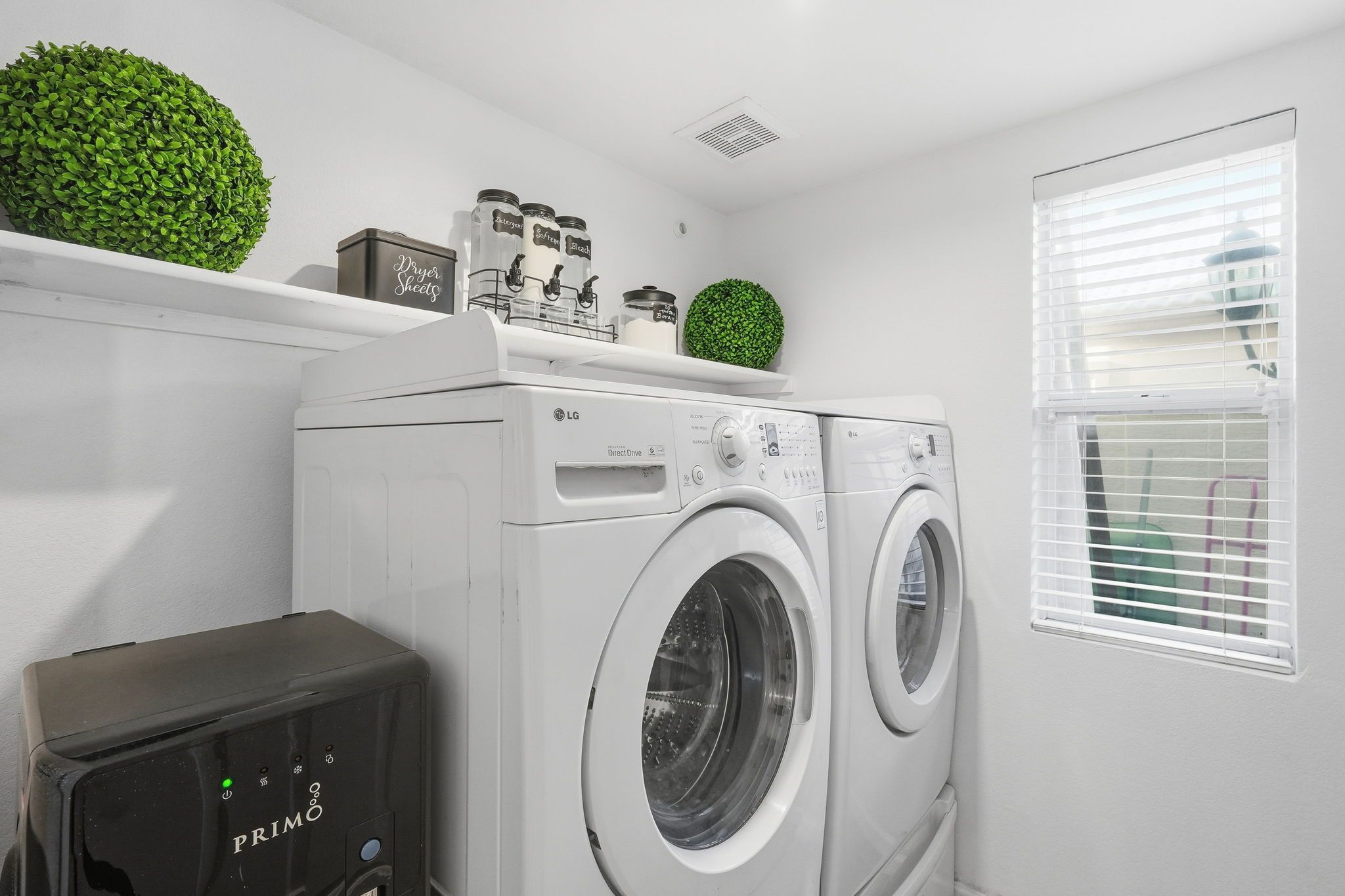A laundry room with two white front-loading washers, a dryer, and a small shelf with jars and a green plant.