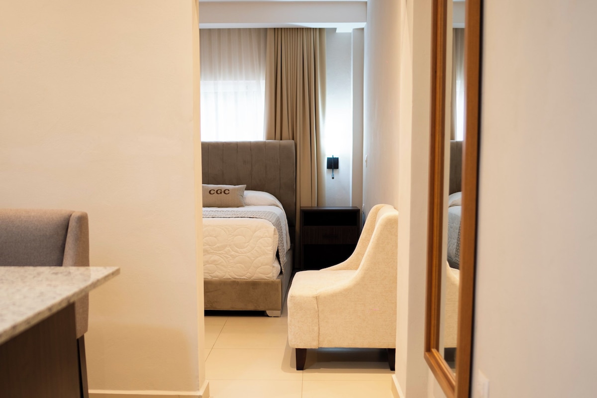 A hotel room with a bed, a seating area with a chair, a desk, and a large mirror on the wall.