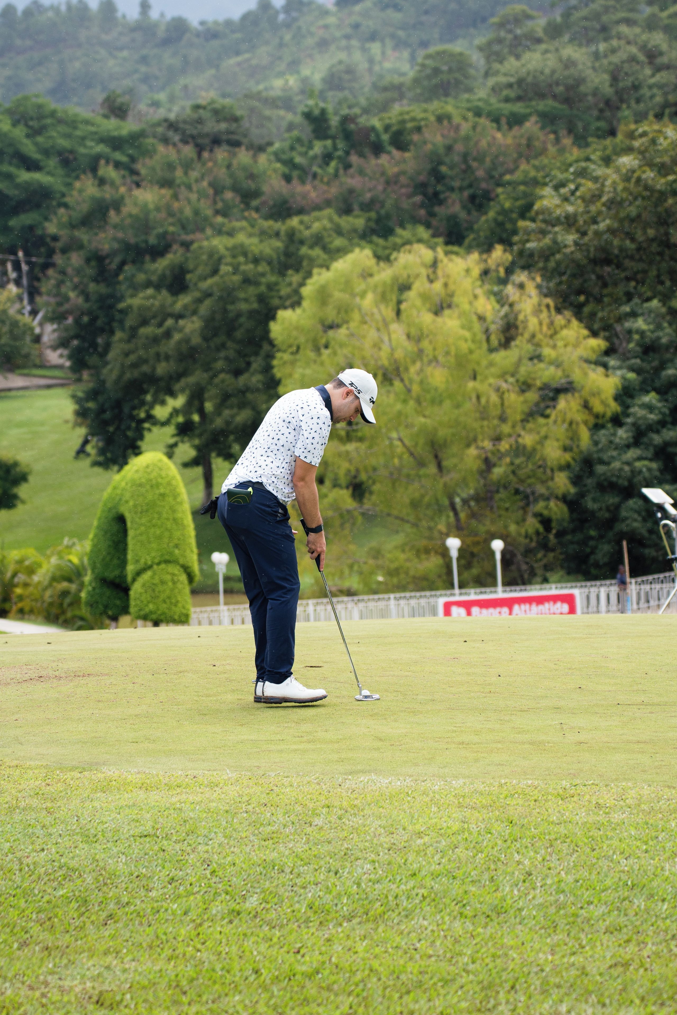Una persona en un campo de golf con un putting green, árboles y una valla de fondo.