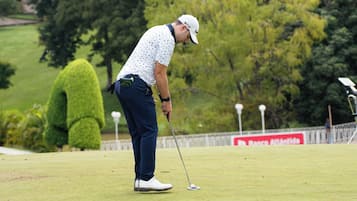 Una persona en un campo de golf con un putting green, árboles y una valla de fondo.