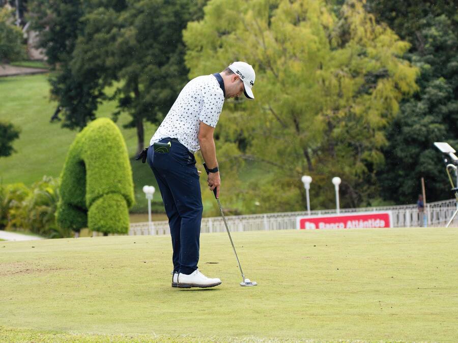 Una persona en un campo de golf con un putting green, árboles y una valla de fondo.