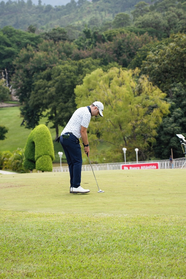 A person on a golf course with a putting green, trees, and a fence in the background.