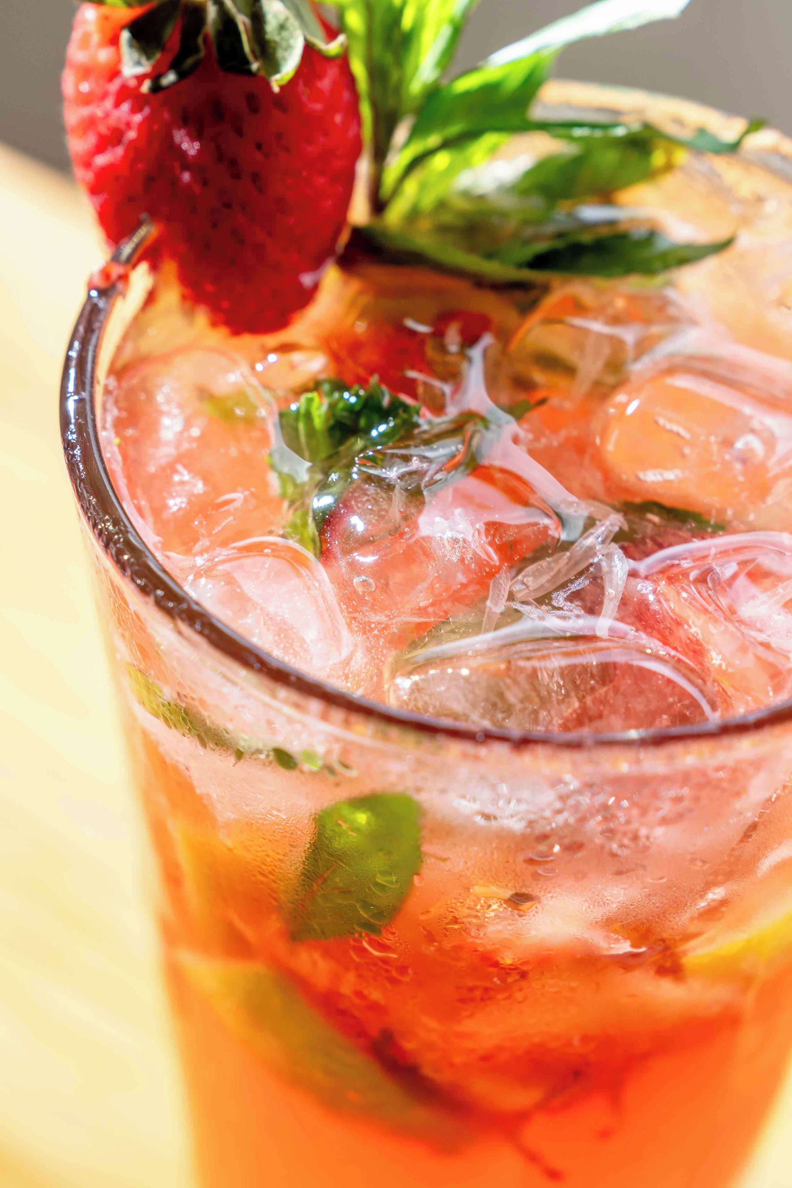 A glass with a red beverage, ice cubes, a strawberry, and mint leaves.