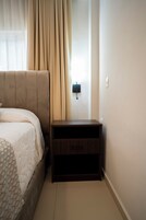 Un dormitorio con cama, mesita de noche, lámpara de pared y ventana con cortinas.