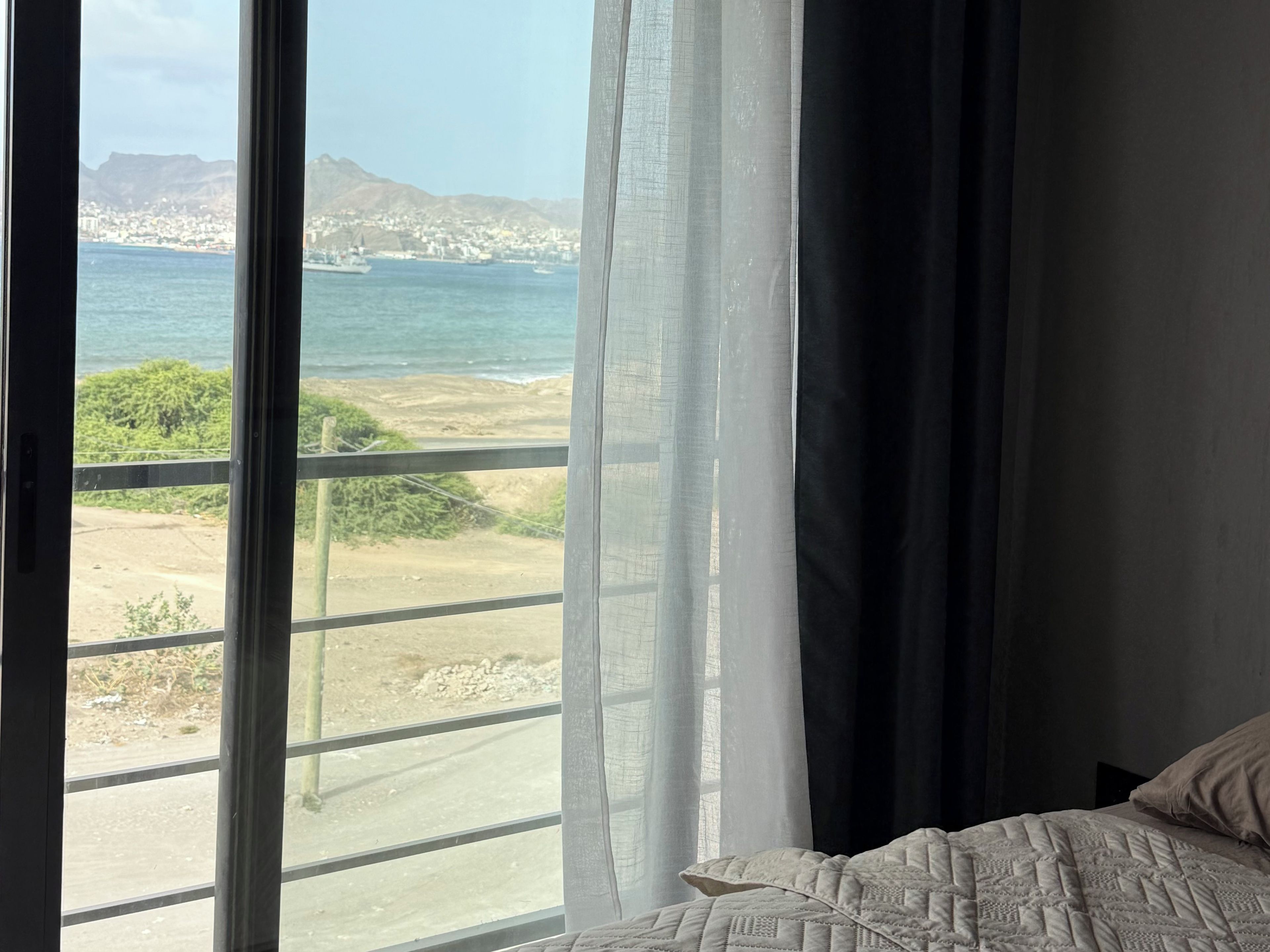Ein Zimmer mit großem Fenster und Blick auf eine Küstenlandschaft, ein Bett mit gemustertem Überwurf und ein dunkler, halb heruntergezogener Vorhang.