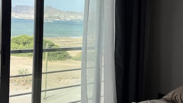 Ein Zimmer mit großem Fenster und Blick auf eine Küstenlandschaft, ein Bett mit gemustertem Überwurf und ein dunkler, halb heruntergezogener Vorhang.