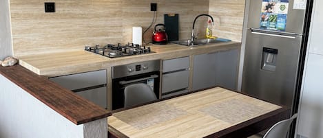 A compact kitchen with a gas stove, a sink, a refrigerator, and a dining table with a chair.