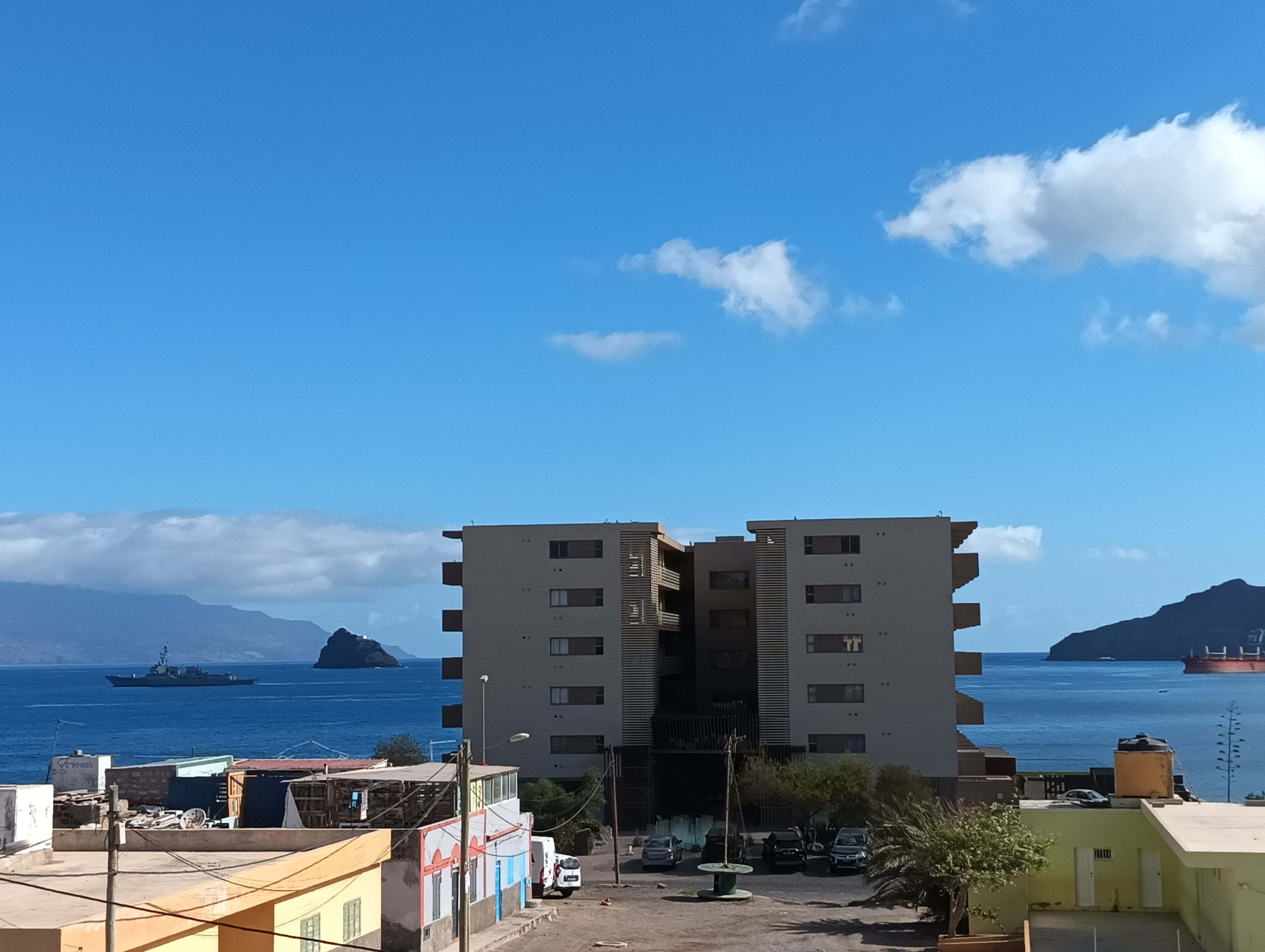 Um edifício de vários andares com telhado plano, situado perto de um corpo de água, com navios visíveis ao longe.
