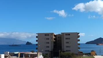 Um edifício de vários andares com telhado plano, situado perto de um corpo de água, com navios visíveis ao longe.