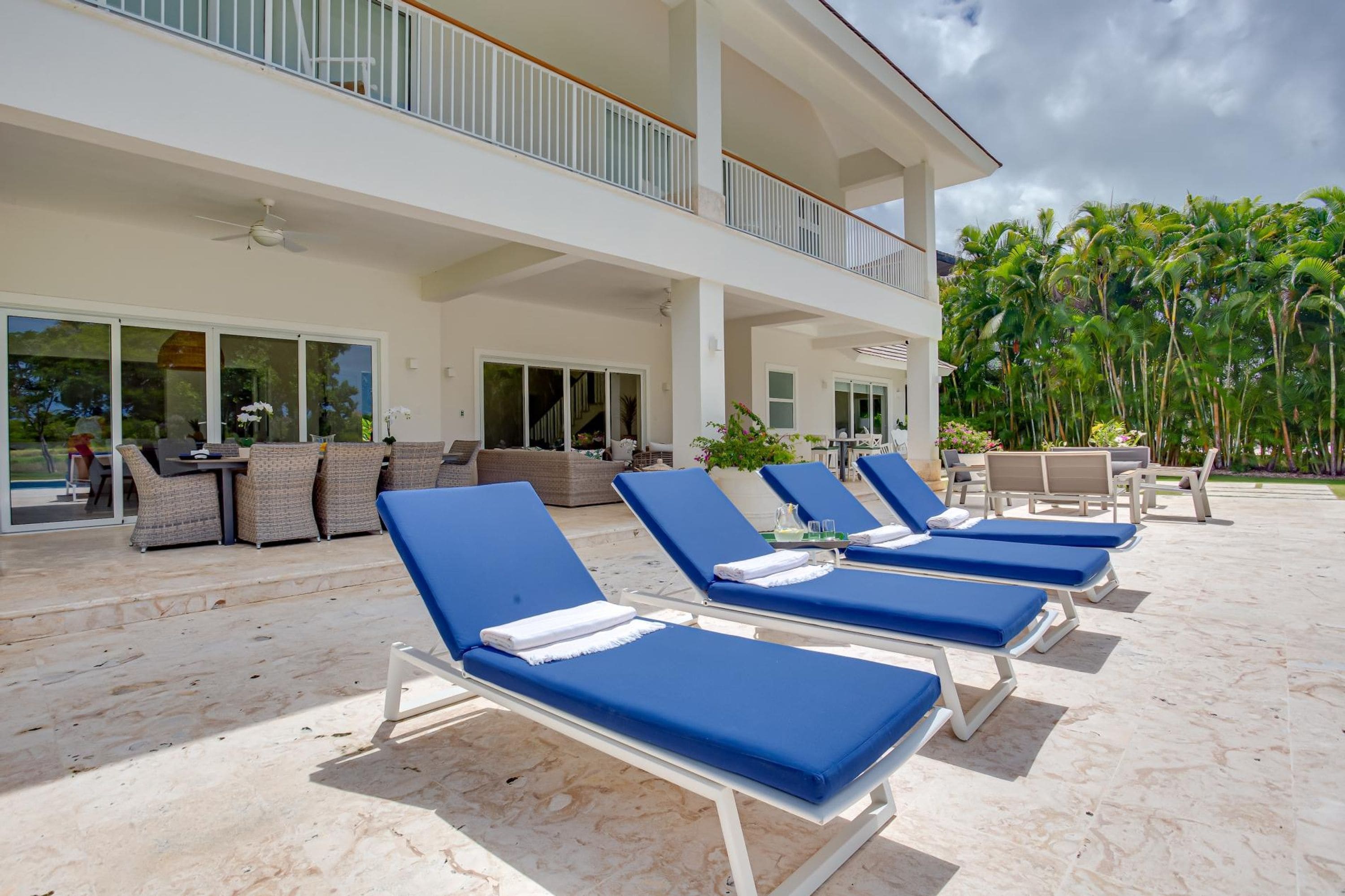 Un resort con zona de piscina, tumbonas, un área de estar cubierta con mobiliario de mimbre y vistas a palmeras.