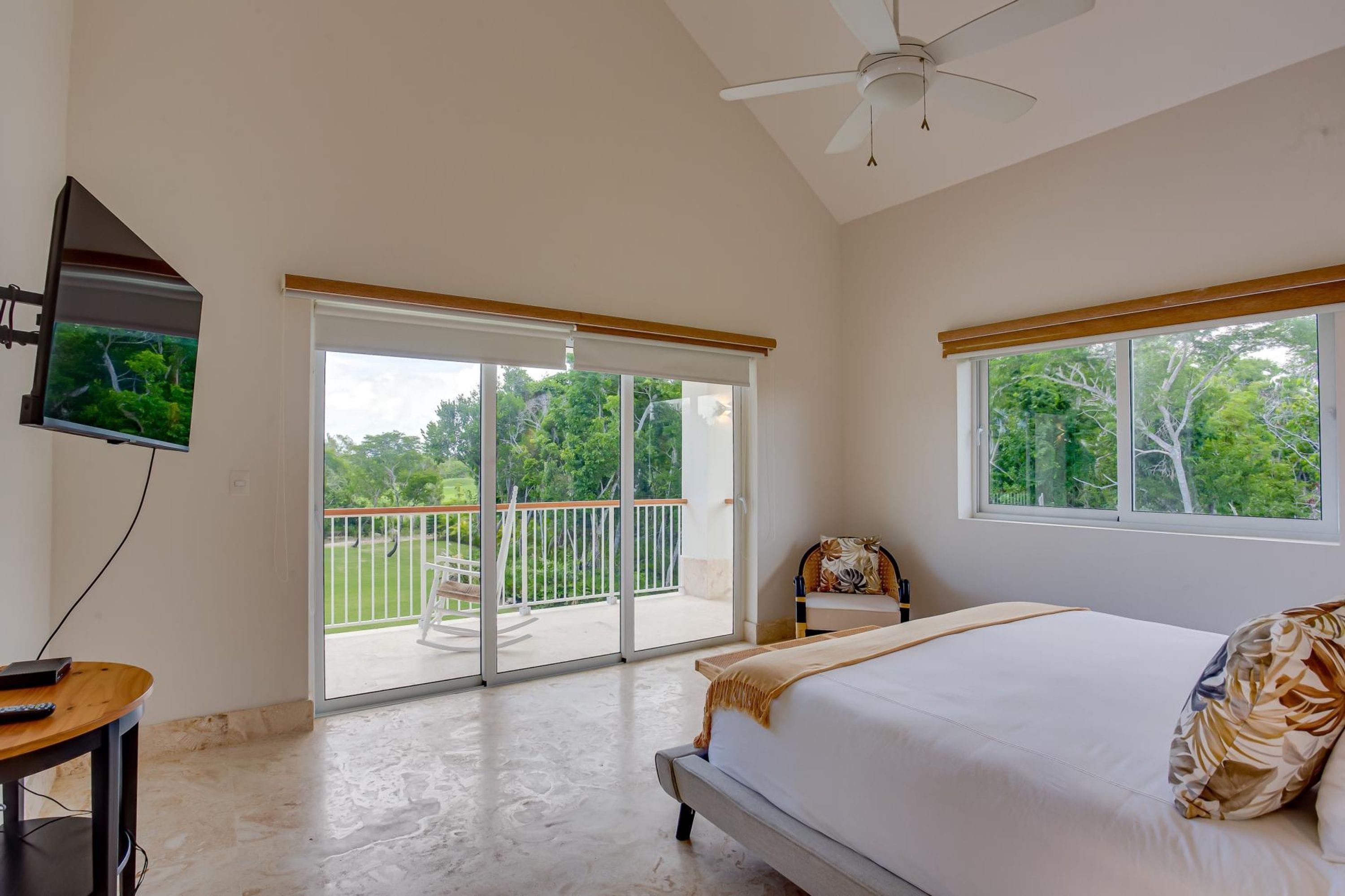 A bedroom with a bed, a television, a ceiling fan, and a balcony with a view of trees.
