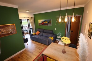 A living room with a sofa, a dining table with chairs, and a green wall with artwork. - Charme e Conforto em Campos: 1º Andar, Elevador e Vaga (Campos do Jordão)