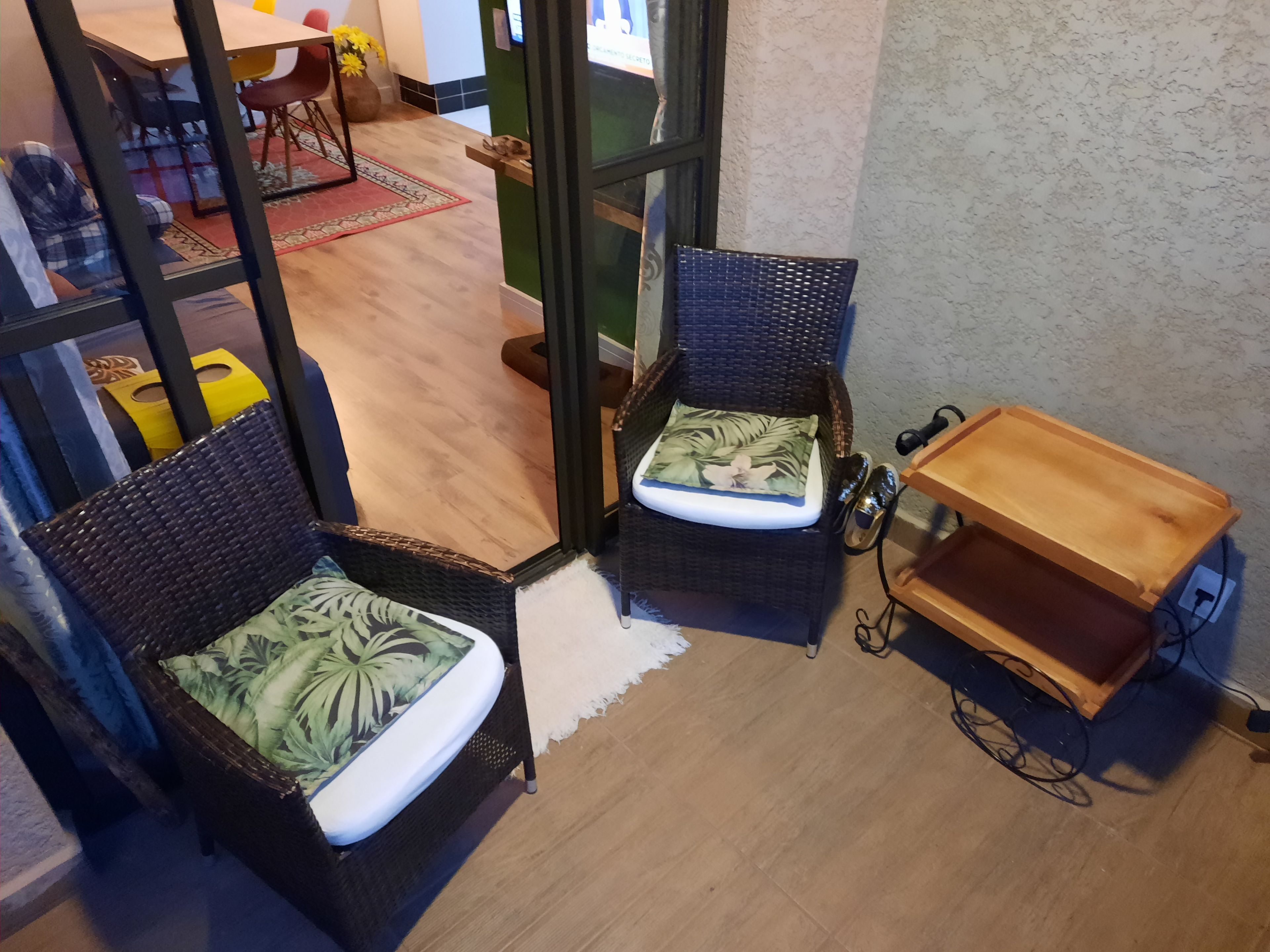 A corner of a room with two wicker chairs with cushions, a wooden side table, and a small shelf with a plant.