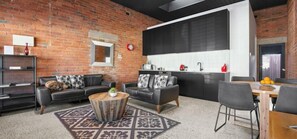 Un espace de vie moderne avec un mur en briques, deux canapés, une table basse et un coin cuisine doté d’un grand tableau noir.