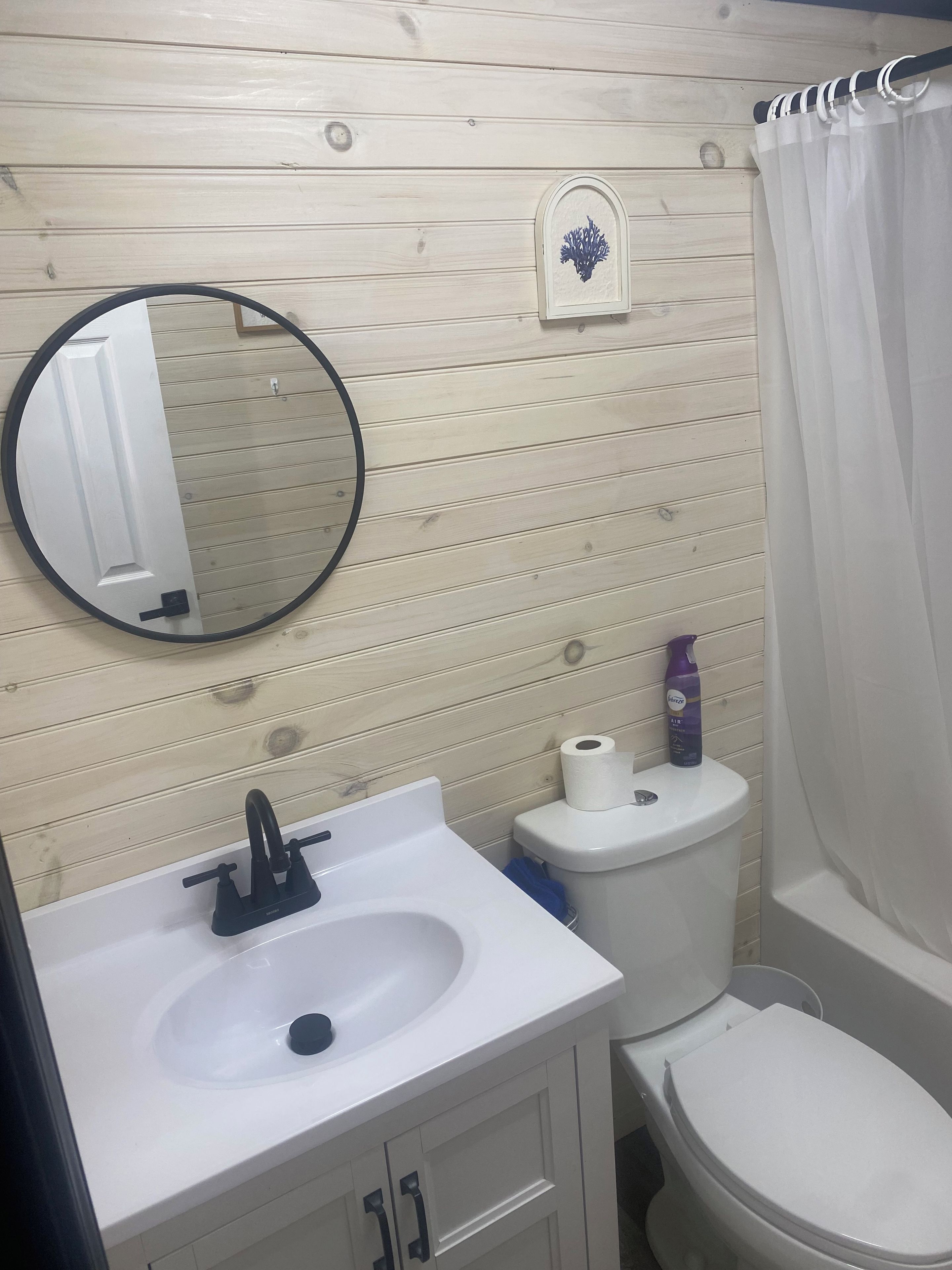 Kylpyhuone, jossa on puinen seinä, pyöreä peili, hana, pesuallas, wc ja suihkuverho.