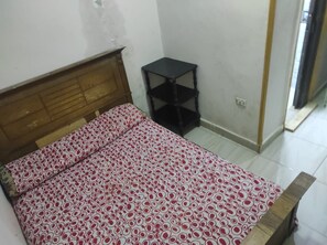 Una habitación con una cama, un pequeño estante negro y una puerta que da a otra habitación.
