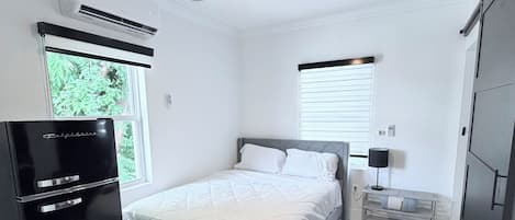 A bedroom with a bed, a chair, a desk with a computer, a ceiling fan, and a window.